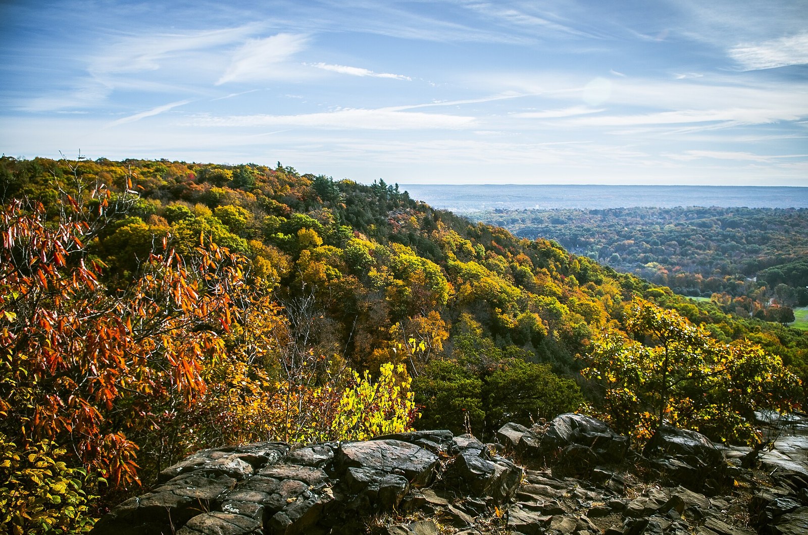 A rocky ridgetop above hillsides of gold, orange and green trees, with a band of water on the horizon.