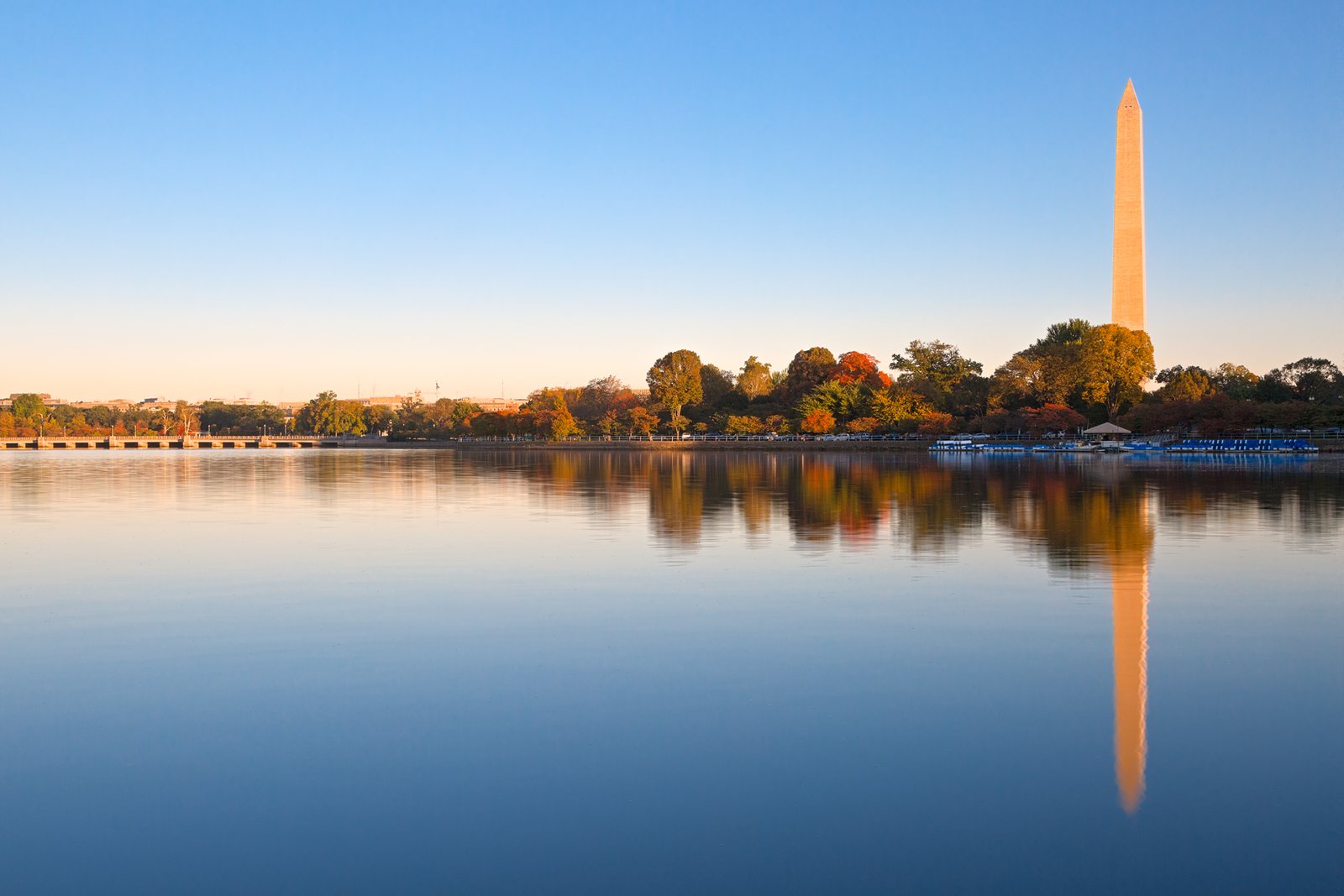 The Washington Monument at sunrise, reflected in the Tidal Basin, with trees along the shore in orange and gold.