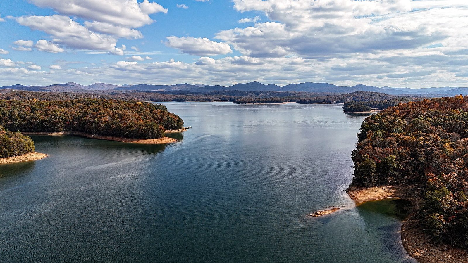 A wide mountain lake with forested shores of orange and brown and blue ridgelines beyond.