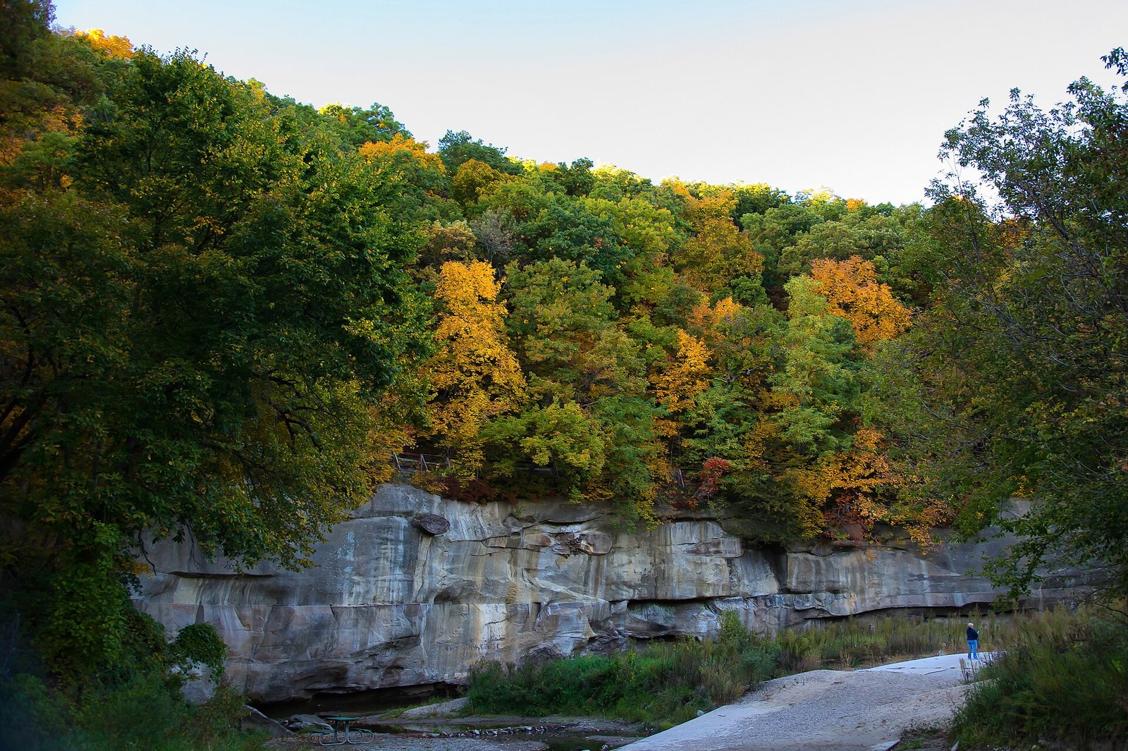 A sandstone cliff beneath trees turning orange and yellow, above a dry creek bed with a hiker in the distance.