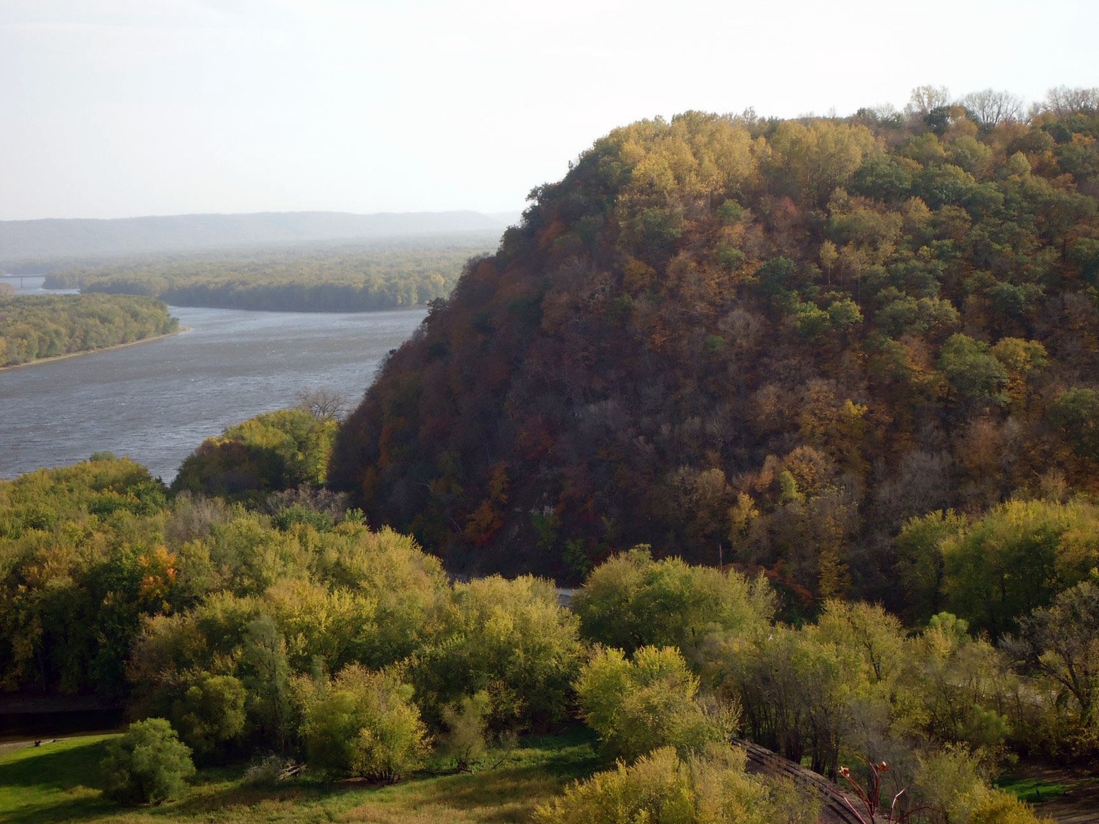 A forested bluff in russet and gold above a wide river with wooded islands under a pale sky.