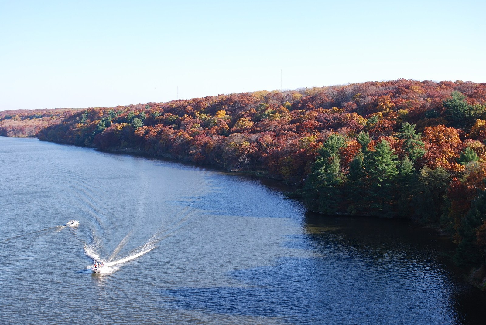 A wooded bluff of red, orange and gold trees above the Illinois River, with two boats on the water.