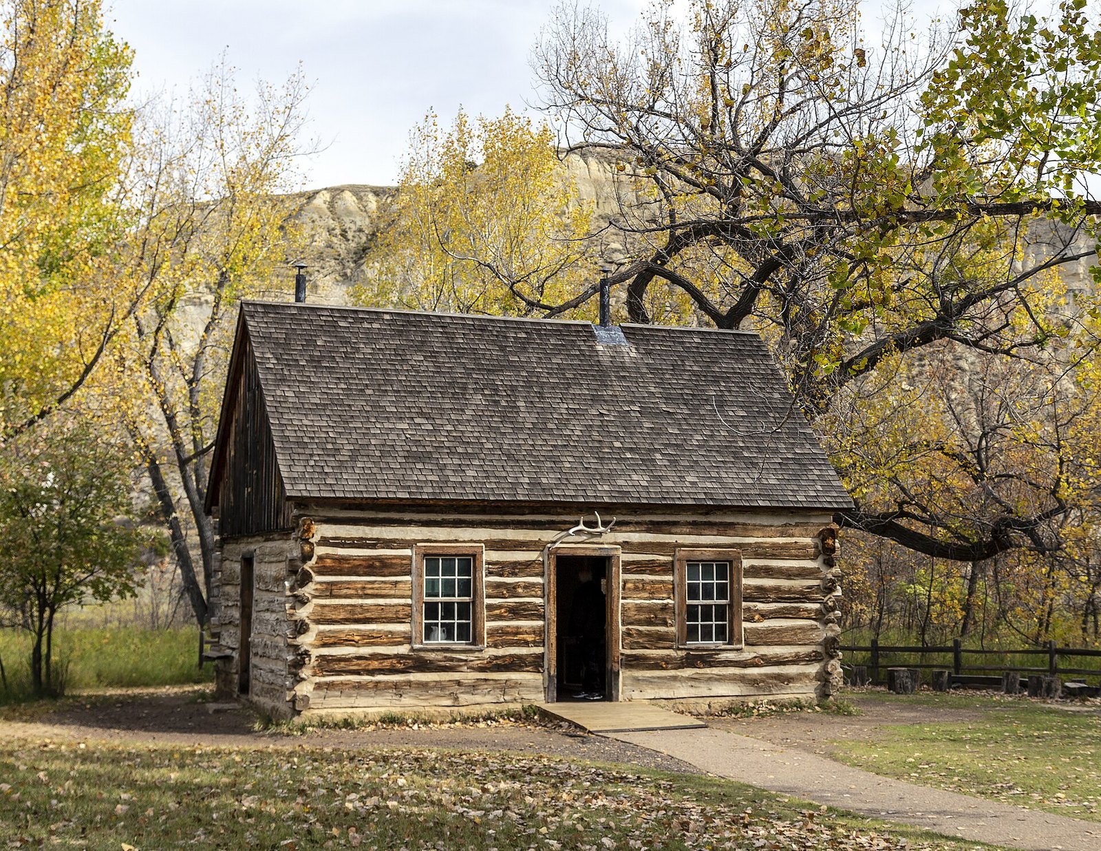 A small log cabin among cottonwoods in yellow leaves, with pale badland bluffs behind.