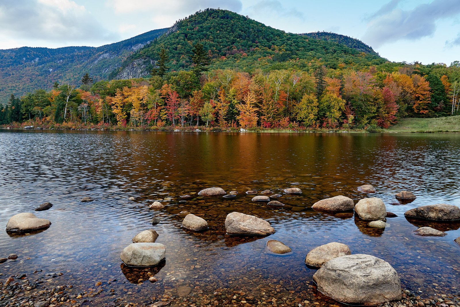 A still reservoir below a rounded green mountain, with the shore trees in red, orange and yellow.