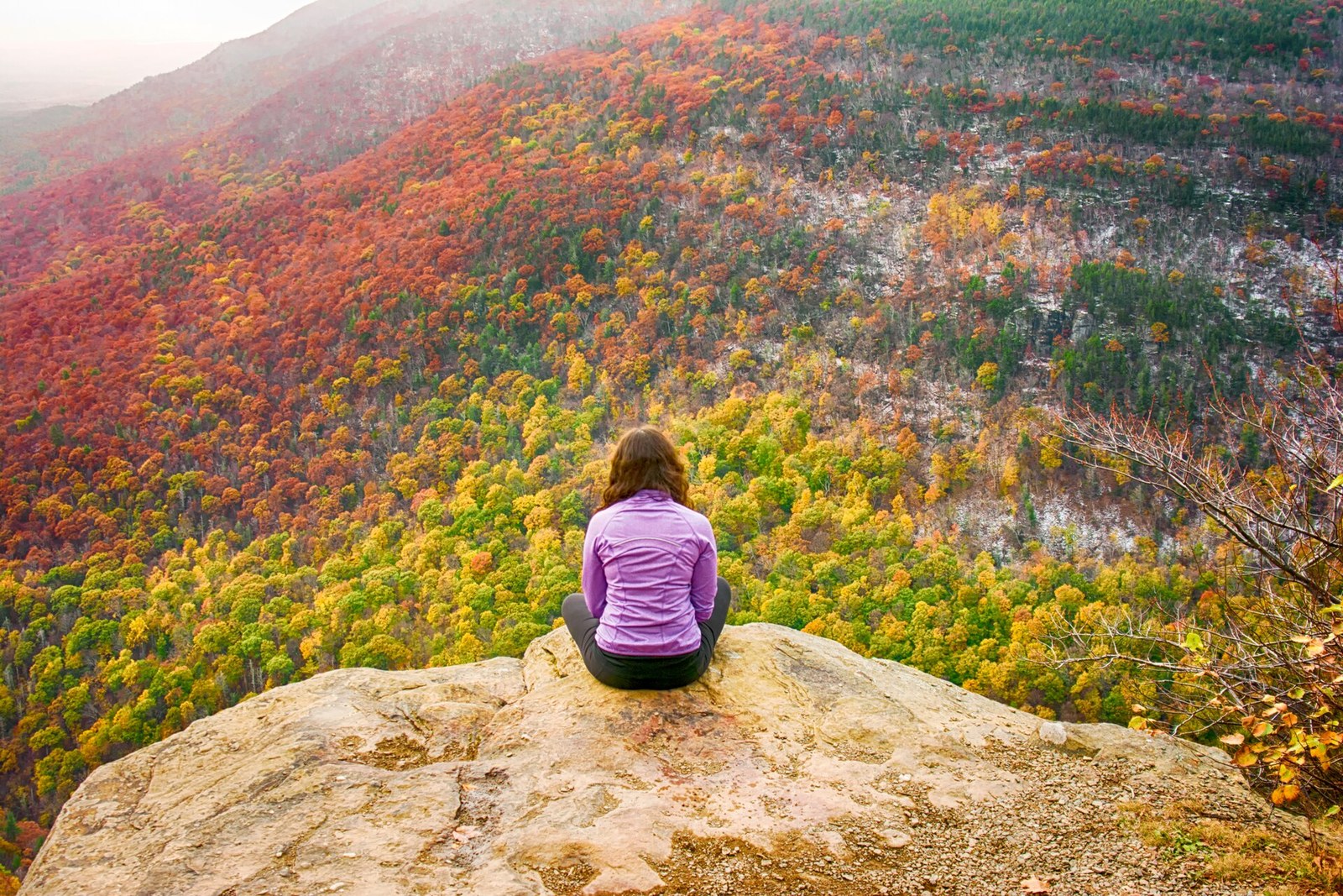 A hiker sitting on a rock ledge above a steep valley covered in red, orange and yellow trees.