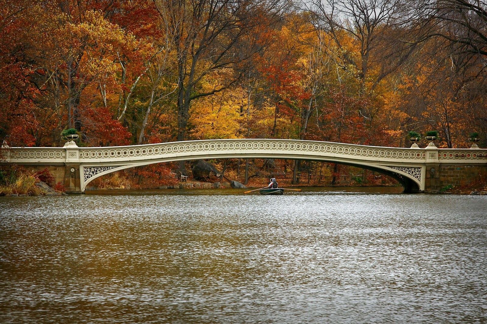 A pale iron bridge arching over a lake, with orange, yellow and red trees behind it.