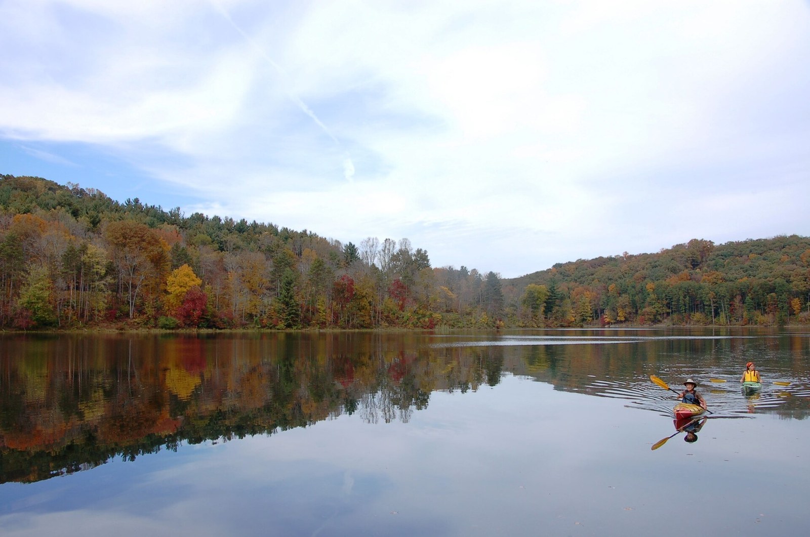 Hills of red, orange and yellow trees reflected in a still lake, with two kayakers on the water.