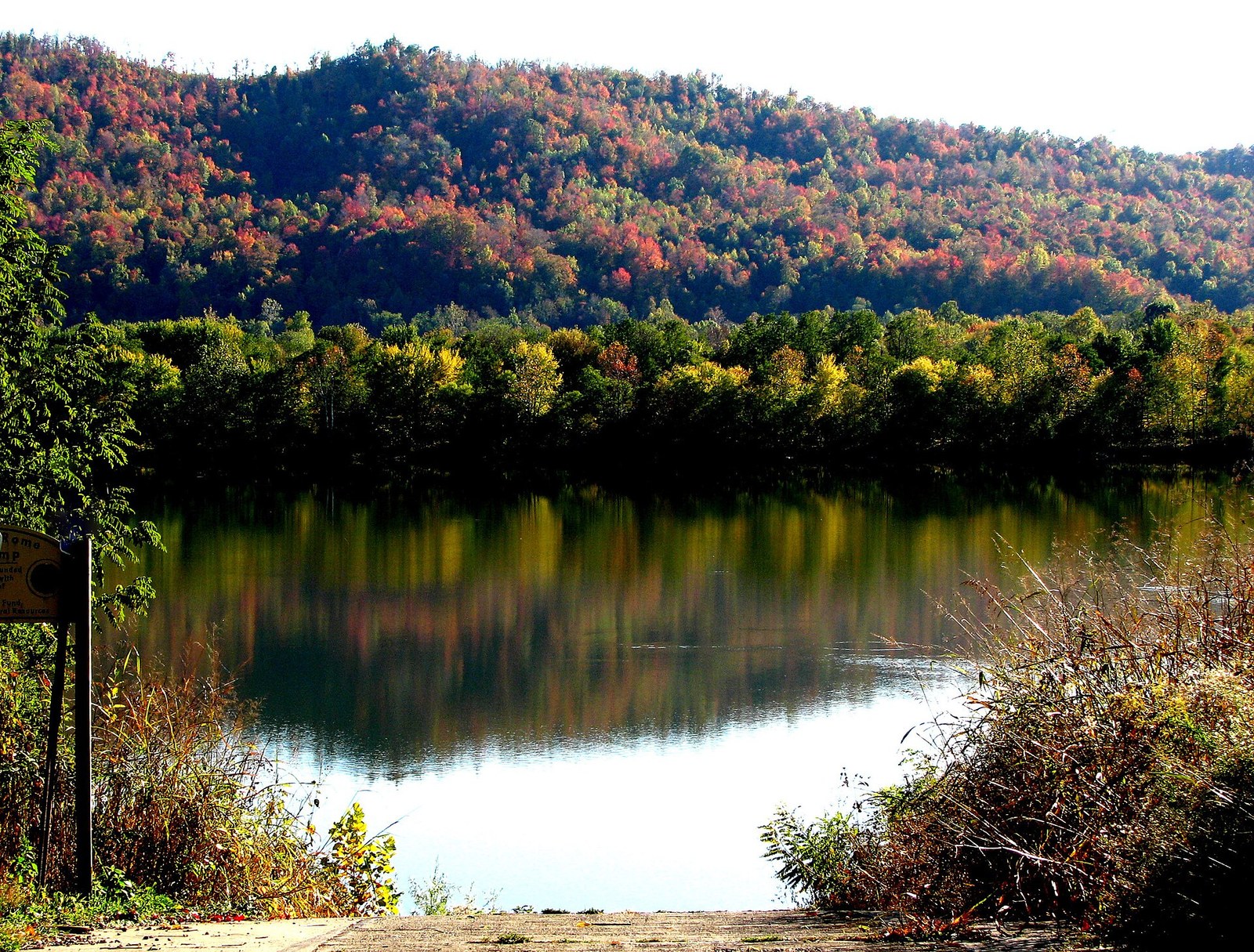 A hillside of red, orange and yellow trees across a calm stretch of the Ohio River, seen from a boat ramp.