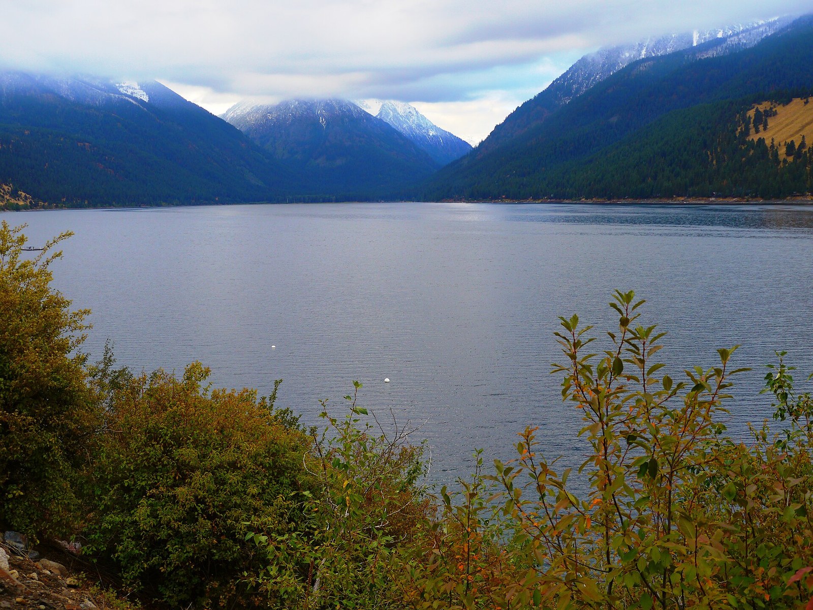 A wide mountain lake below snow-dusted peaks, with shrubs in orange and yellow leaves along the near shore.