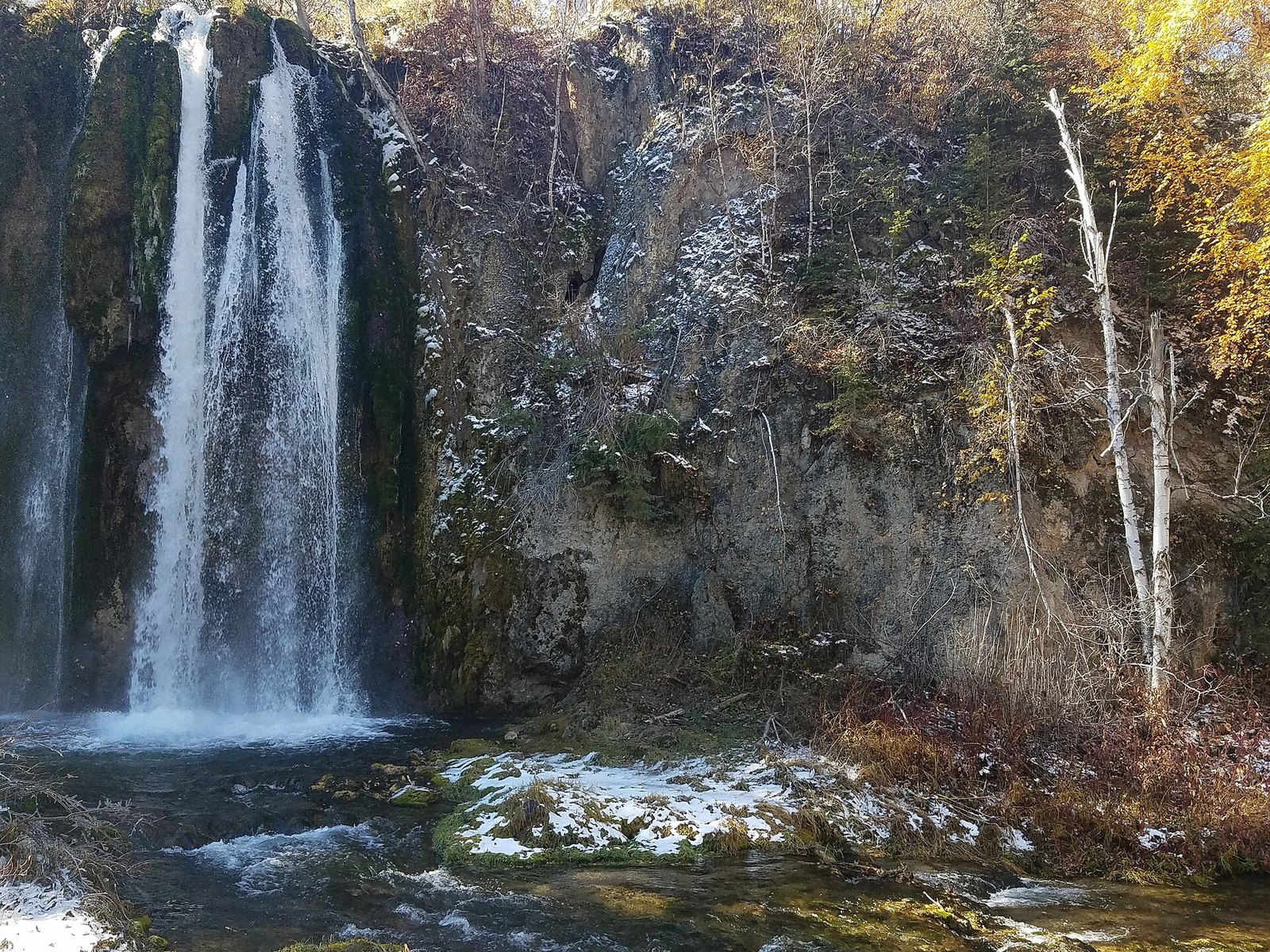 A tall waterfall dropping down a dark cliff dusted with snow, with yellow leaves at the upper right.