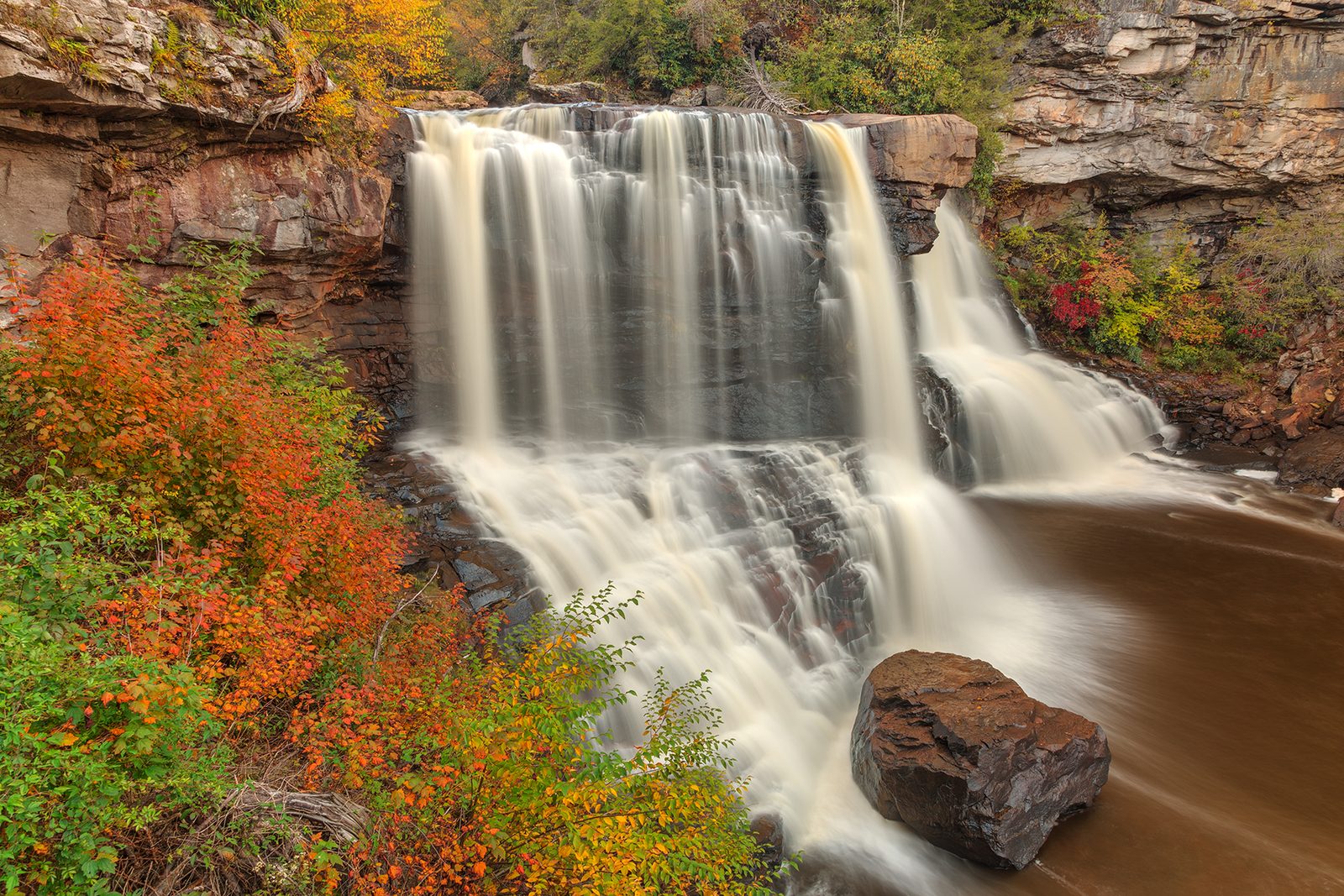Blackwater Falls in a long exposure, with orange and red shrubs along the cliff beside it.