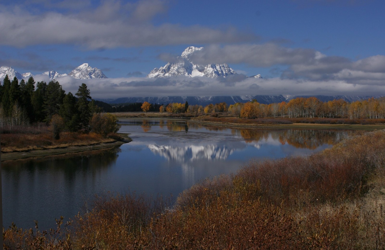 A calm river bend reflecting snow-covered peaks, with orange and gold trees on the far bank and brown shrubs in the foreground.